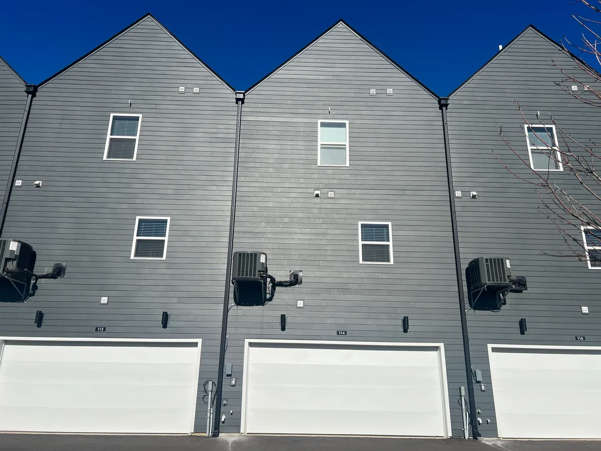 A contemporary exterior view of the property showcasing gray siding, crisp white garage doors, and neatly arranged windows, with visible air conditioning units. The organized, modern facade hints at a well-maintained residence in a lively neighborhood setting.