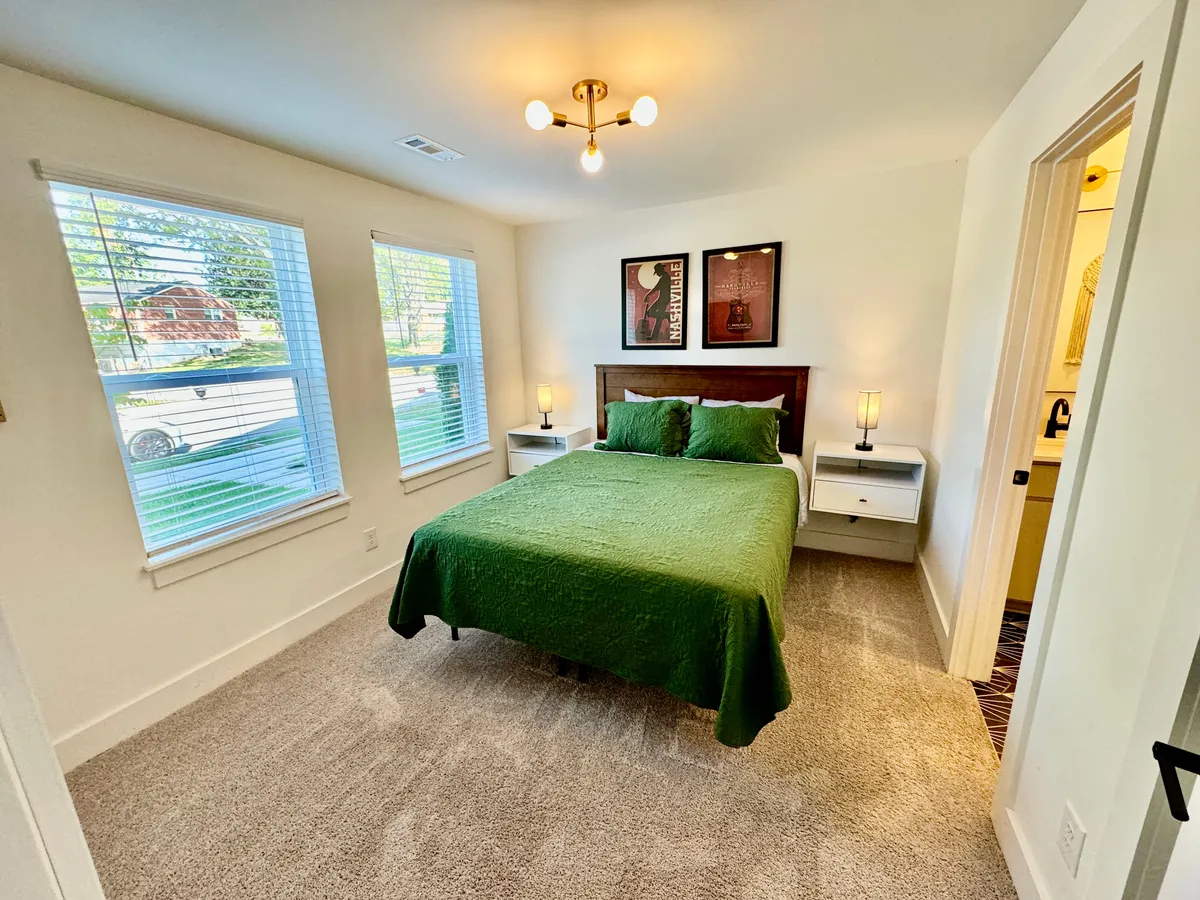 A cozy bedroom with a bed dressed in green bedding, two bedside lamps for soft lighting, framed art above the bed, and windows letting in morning light. The room exudes comfort, privacy, and a sense of peacefulness.