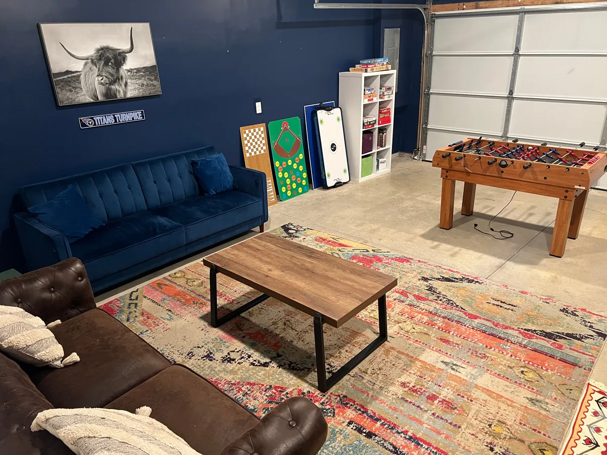 A spacious interior recreation area featuring a blue couch, foosball table, coffee table, and a colorful rug. Wall art and a TV suggest a setting for relaxation and fun activities, ideal for groups.