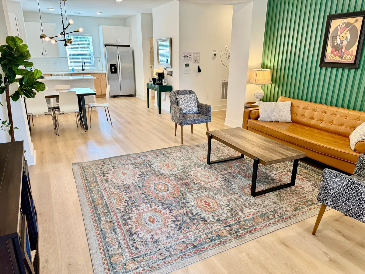 An open-plan dining and living area with a green accent wall, tan leather sofa, area rug, and a dining table. The space is brightly lit, welcoming, and stylish, perfect for gatherings.