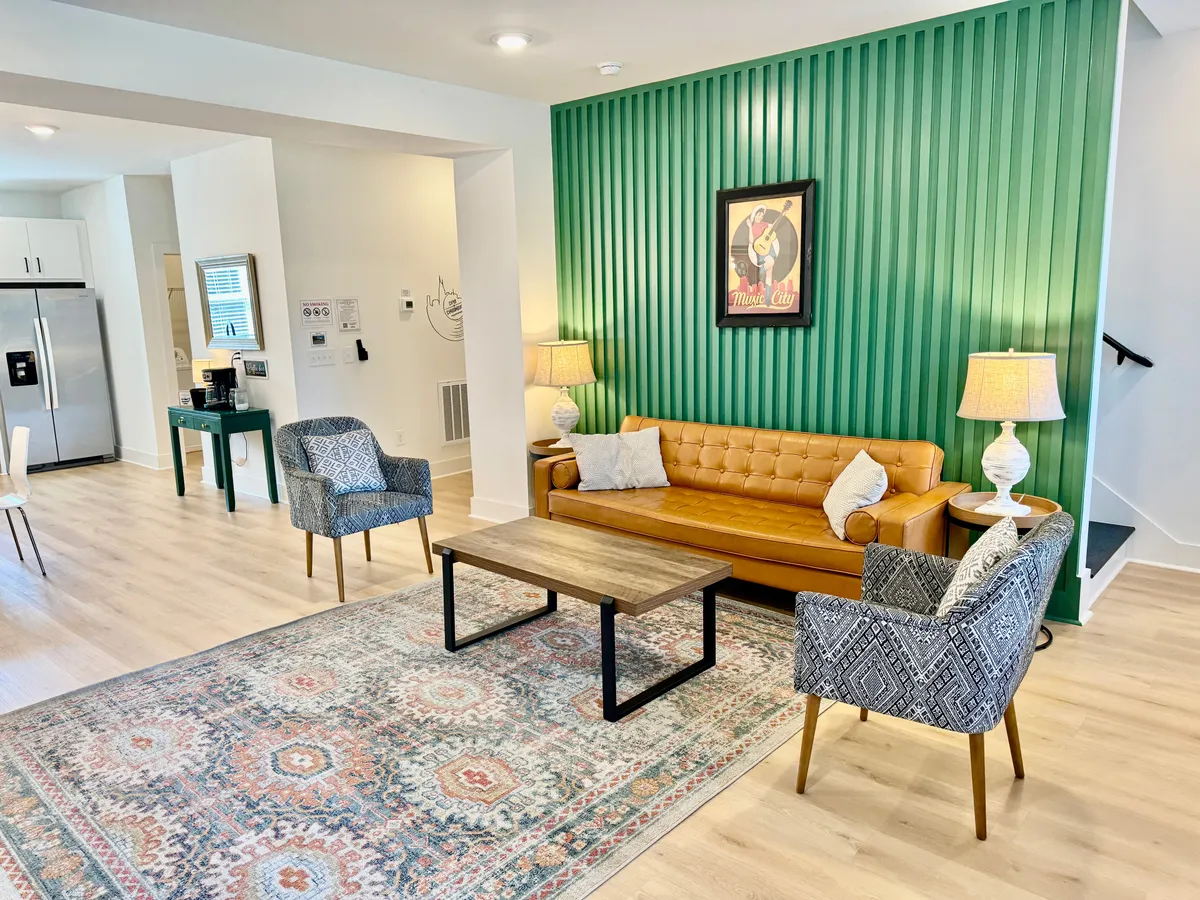 The image showcases a stylish, welcoming living space featuring a rich green accent wall, a plush tan leather sofa, a vibrant colorful rug, a sturdy wood coffee table, and decorative modern lamps. The room is designed for comfort and relaxation, perfect for gathering with friends or family for a cozy evening in Nashville.