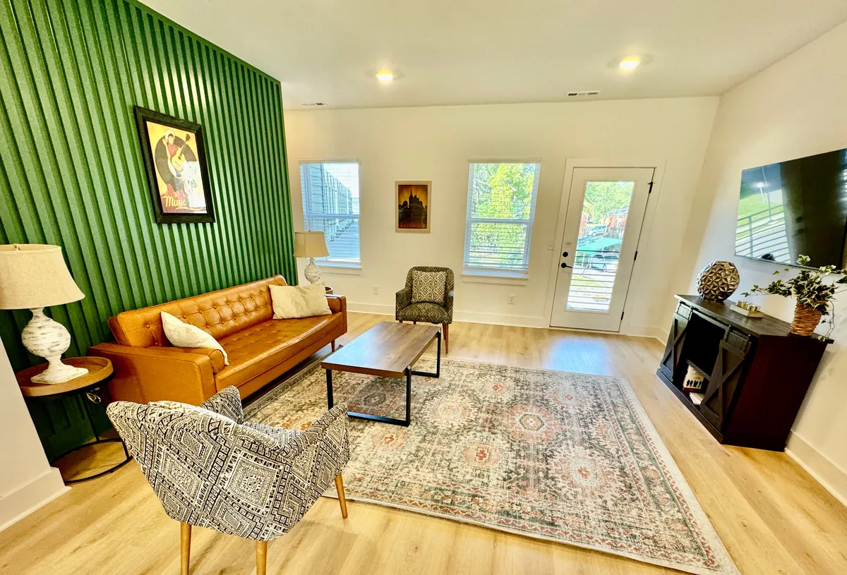 A stylish and welcoming living room featuring a green accent wall, brown leather sofa, decorative coffee table, area rug, and a modern TV unit. The space is airy with plenty of natural light and is arranged for both relaxation and social gatherings.