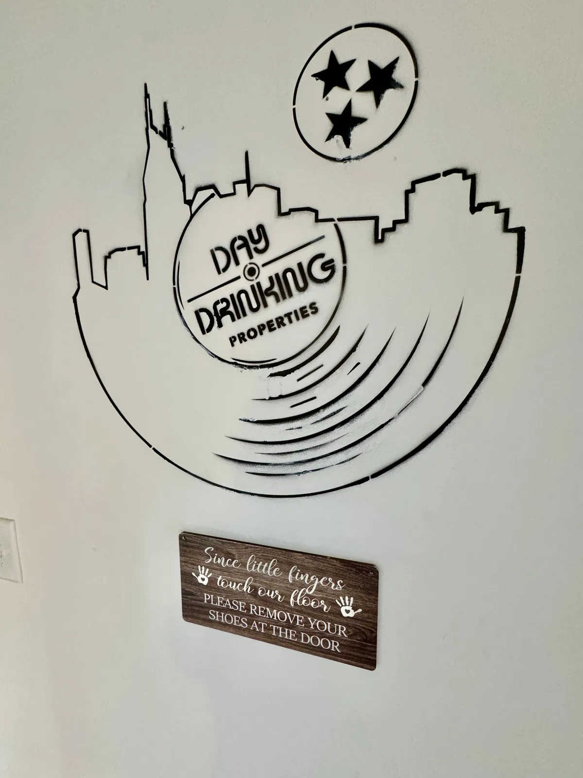 A playful entry area displaying the Day Drinking Properties logo, a Nashville skyline silhouette, and a welcoming shoe removal sign. The scene exudes a sense of home and hospitality.