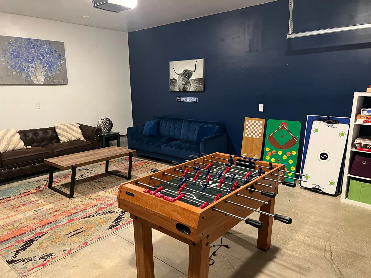 This image captures a vibrant garage game room setup featuring a foosball table, a blue couch for lounging, a coffee table, and colorful wall art. The space is designed for play, relaxation, and hosting lively gatherings, exuding an energetic and friendly atmosphere.