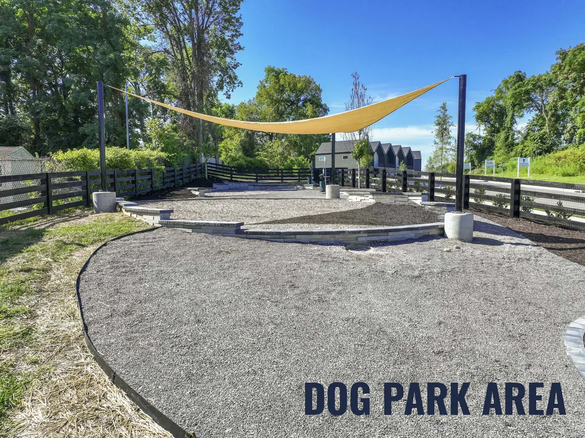 The image showcases a cheerful community dog park with a shaded area, a gravel surface for pups to run and play, and people socializing with their dogs. The park is inviting and relaxed, encouraging both pets and their owners to mingle and unwind.